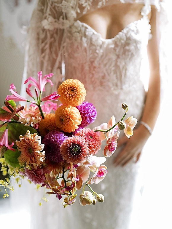 Bridal bouquet, colorful bridal bouquet of orchids, dahlias, and lilies held against a strapless lace wedding dress in window light