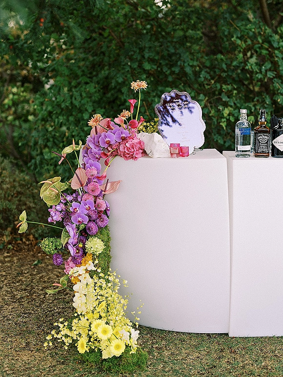 Wedding bar setup with a white bar counter, orchid floral installation, and votive candles, set on a garden lawn with greenery backdrop