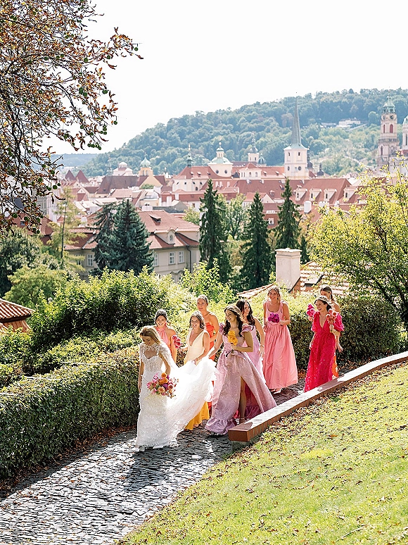 Bridesmaid group walking with the bride along a cobblestone path, carrying bouquets in colorful dresses with veil, skyline beyond hedges