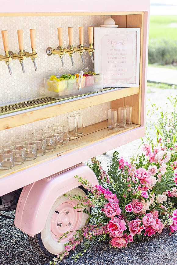 Mobile bar setup with drink taps, cocktail menu sign, and citrus garnish display on wooden shelves against a pink trailer on gravel