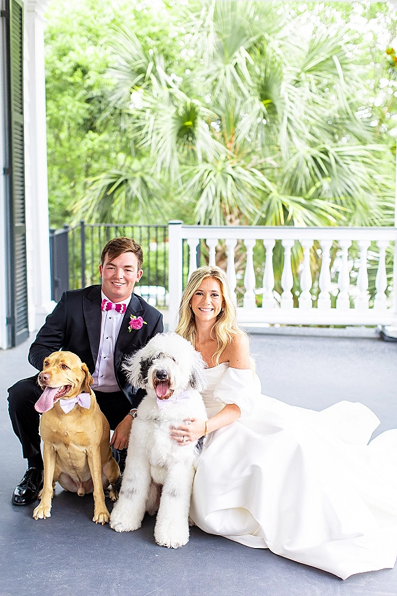 Couple portrait of newlyweds with dogs on a porch, bride in off-shoulder gown and groom in tuxedo, dogs in bow ties by white railing and palms