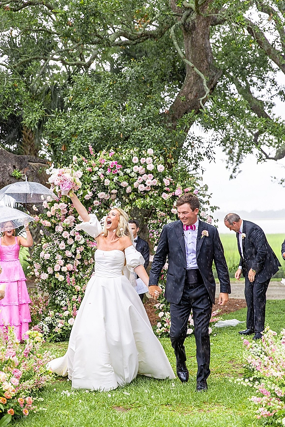 Wedding recessional as bride and groom walk out holding hands under clear umbrellas, rose petals flying on a rainy garden lawn