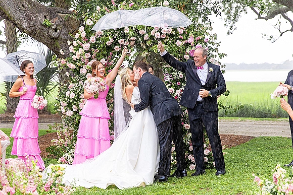 Ceremony kiss under clear umbrellas in a rainy wedding ceremony, bride in long veil and groom in black suit beneath a rose arch on grass