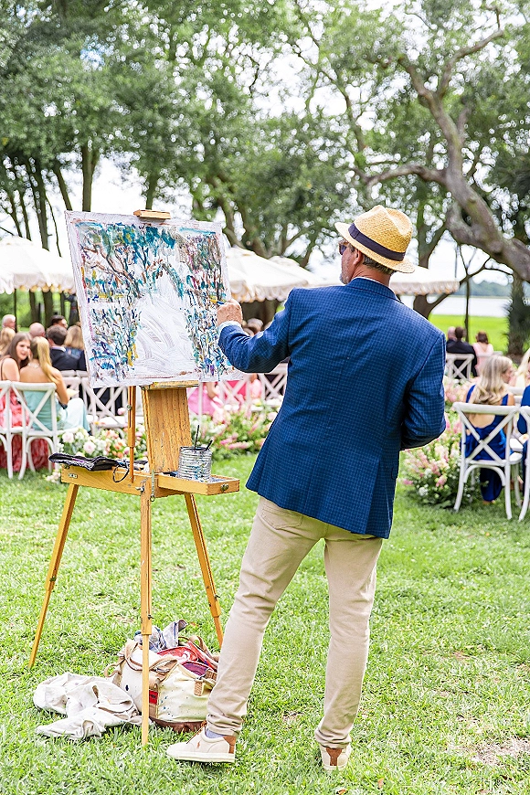 Wedding live painter creating a live wedding painting on an easel with paint palette under a tent canopy at a lakeside ceremony lawn