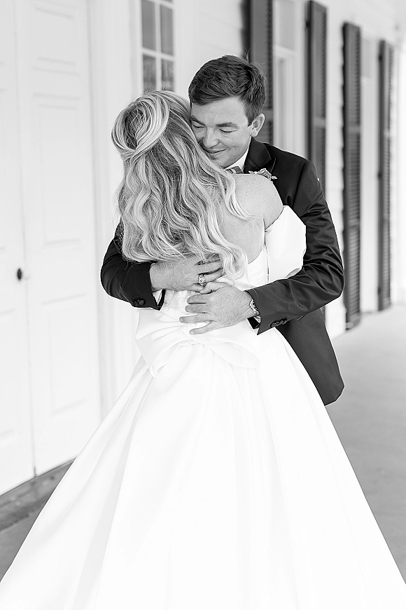 First look moment as the groom hugging bride in a black tuxedo, holding her strapless satin ball gown on a porch by white windows