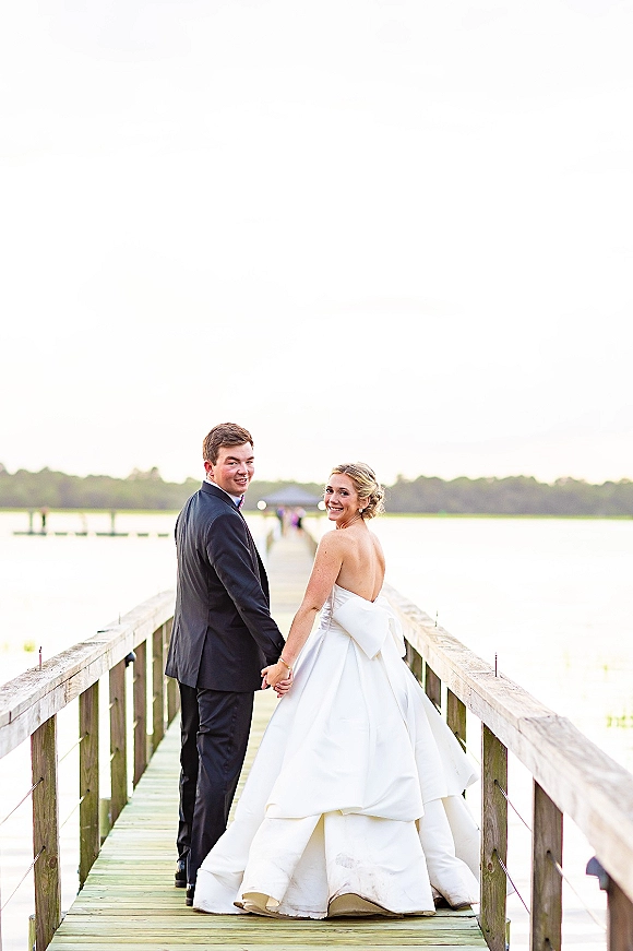 Couple portrait of bride and groom holding hands on a wooden dock by a lake, bride in strapless gown with oversized bow looking back