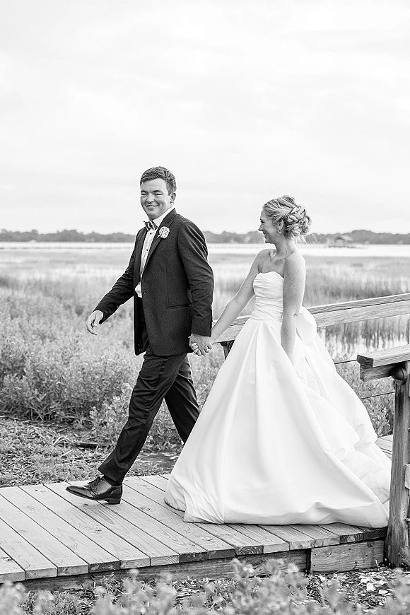 Couple portrait of bride and groom walking hand in hand on a wooden boardwalk by marsh grass and water, bride in strapless gown