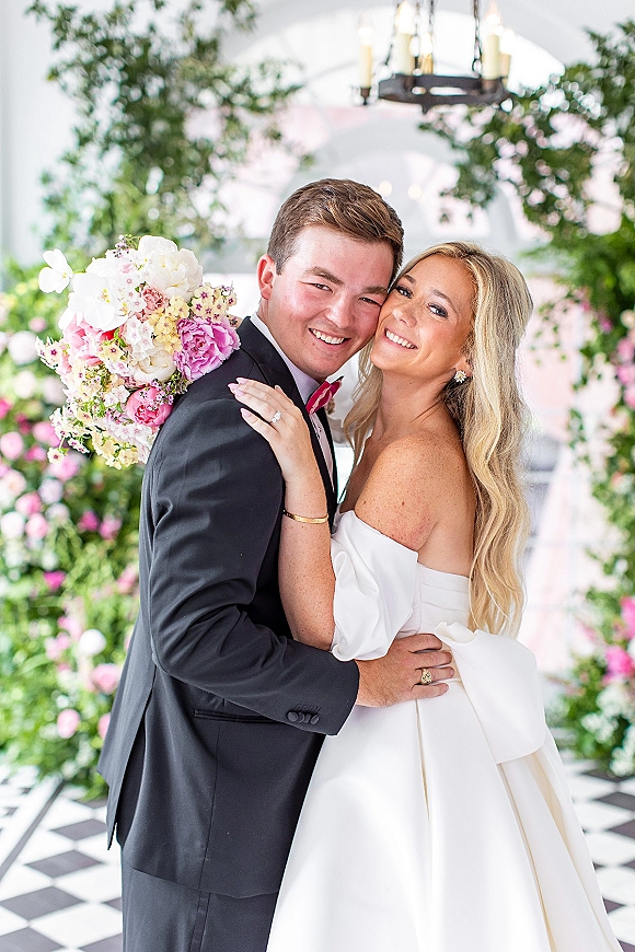 Couple portrait of bride and groom hug under a floral arch, bride holding peony bouquet beside a chandelier in a glass-ceiling conservatory