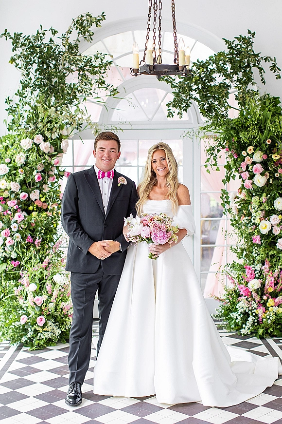 Couple portrait of bride and groom posing under a floral arch, bride holding bouquet in an indoor conservatory with arched window and chandelier