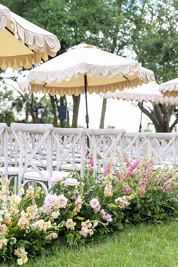 Outdoor ceremony seating with garden wedding ceremony chairs in neat rows, cream umbrellas overhead, and pastel meadow flowers lining the grassy aisle under trees