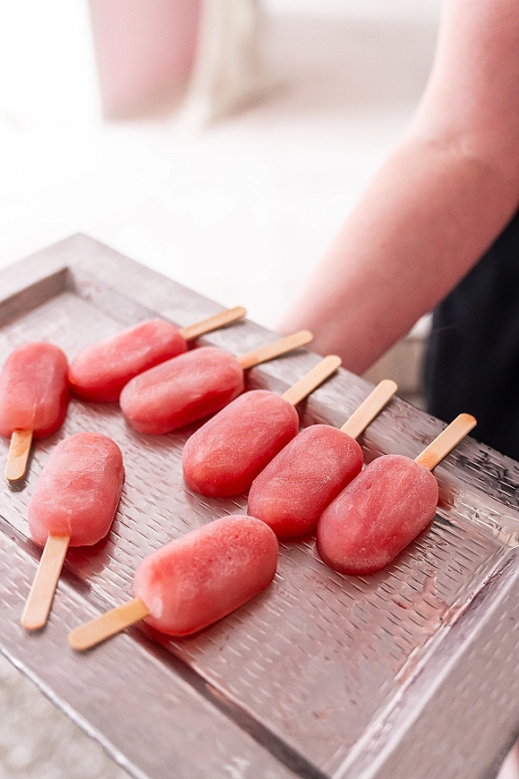 Wedding popsicles on wooden sticks in a serving tray, popsicle wedding favors in pink tones held by a person in bright indoor light
