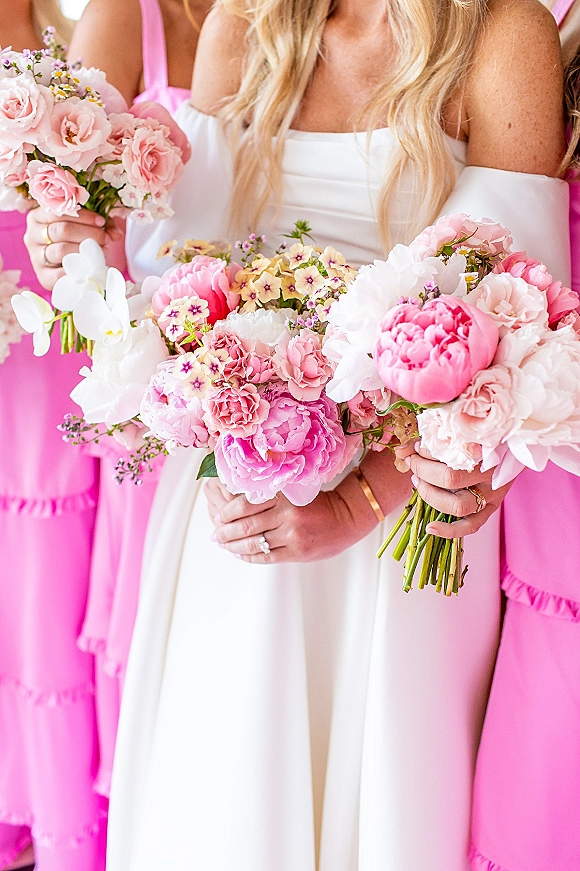 Bridesmaid bouquets with pink peony blooms, roses, and white orchids held against bright pink dresses indoors, with lush greenery accents