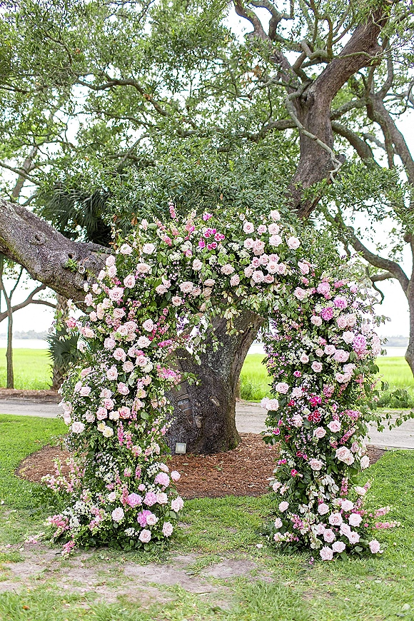 Floral ceremony arch with rose florals and greenery on a lawn beneath an oak tree, framing a dirt path in an open field