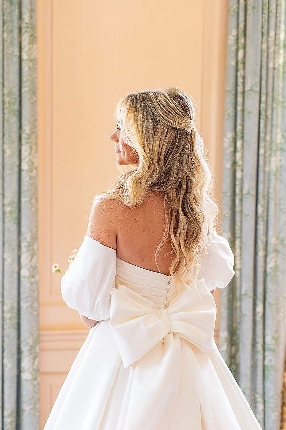 Bridal portrait of a bride in a backless wedding dress with fabric-covered buttons and an oversized bow, wavy blonde hair, indoors by paneled wall