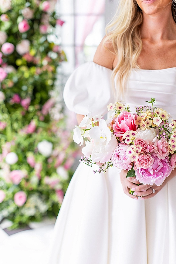 Bridal portrait of bride holding bouquet of pink peonies and white orchids, wearing an off the shoulder gown before a floral wall indoors