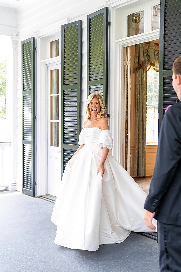 Bride portrait in an off the shoulder wedding dress with puff sleeves, holding her skirt on a white porch by a doorway with green shutters
