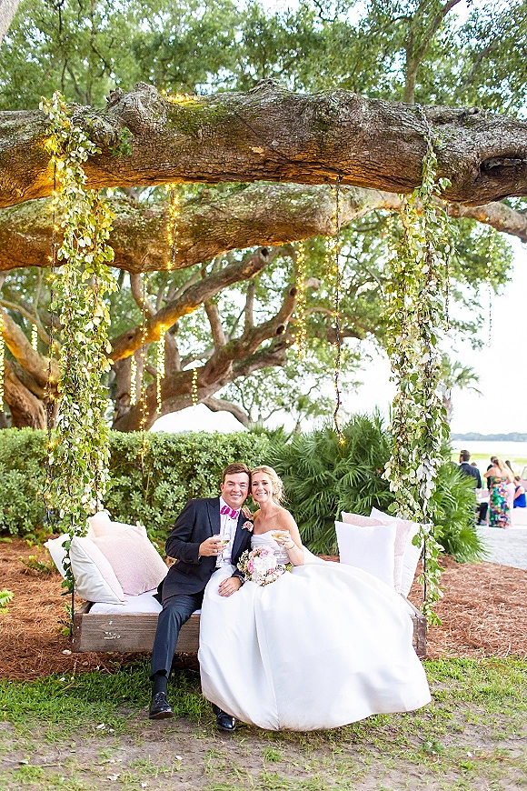 Couple portrait of bride and groom on swing, in strapless wedding dress and tuxedo, holding champagne under string lights by waterfront greenery