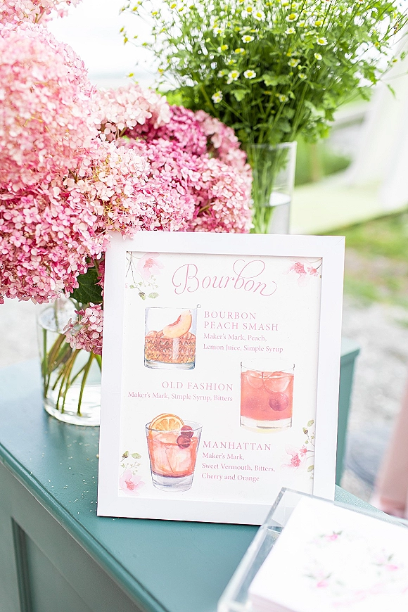 Wedding cocktail menu signature drinks sign in a framed card with watercolor illustrations and pink hydrangeas on a green table outdoors