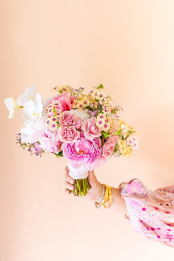 Bridal bouquet of pink peonies and roses with white orchid and greenery stems, held by an embroidered sleeve against a peach backdrop