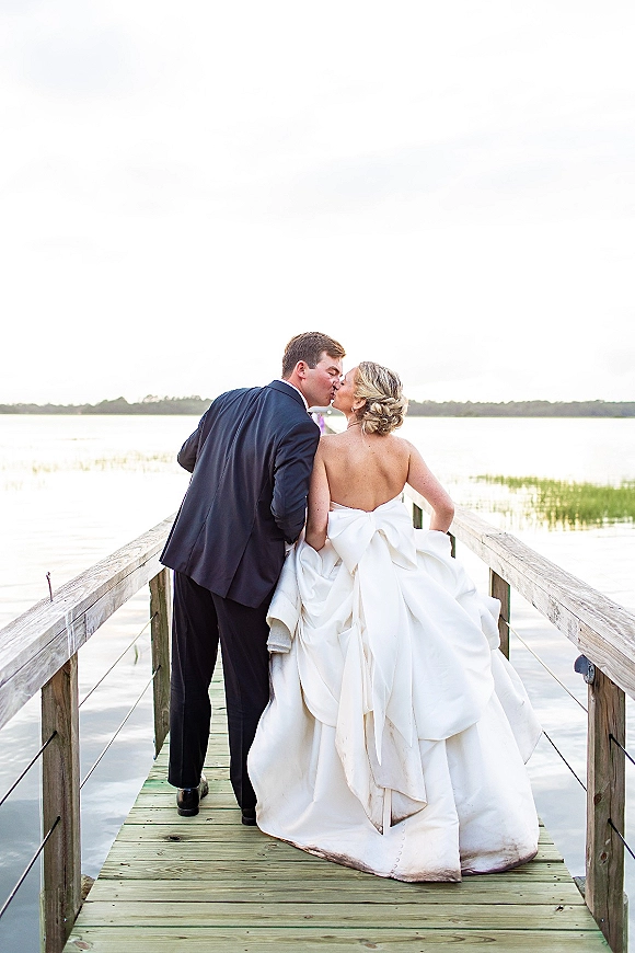 Wedding kiss portrait of bride and groom kissing on a wooden dock, bride in strapless gown with oversized bow back by the lake shoreline