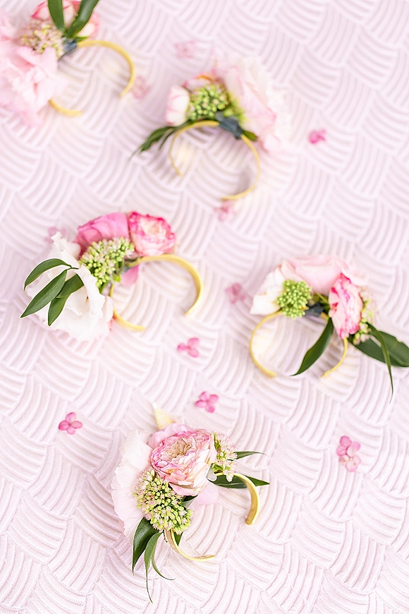 Wedding boutonniere with pink boutonniere blooms and greenery, wrapped on a gold hoop, on textured pale pink fabric with scattered petals