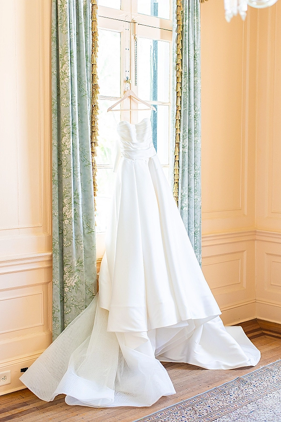 Wedding dress with a strapless sweetheart neckline hanging on a hanger in a tall window, full skirt and train framed by curtains