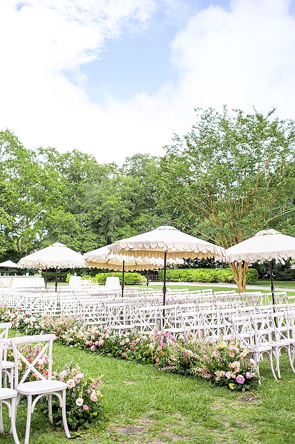 Outdoor ceremony setup with garden wedding ceremony seating, white folding chairs under patio umbrellas and floral-lined aisle on a lawn