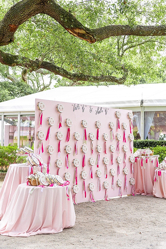 Wedding escort display with a wedding escort card wall of hanging embroidery hoops and pink ribbons against a pink backdrop under a tented outdoor setting