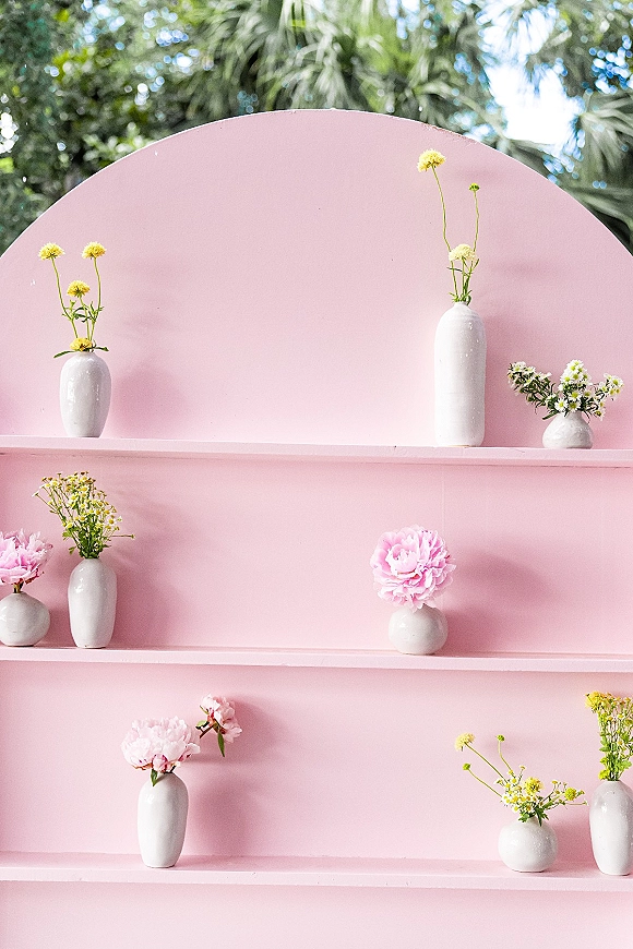 Floral backdrop decor with a pink wedding backdrop arch, display shelves of white ceramic vases and peony blooms in outdoor greenery