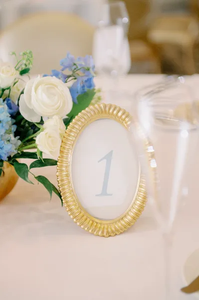 Wedding table number in a gold frame with white roses, blue flowers, and greenery on a white tablecloth beside a water glass