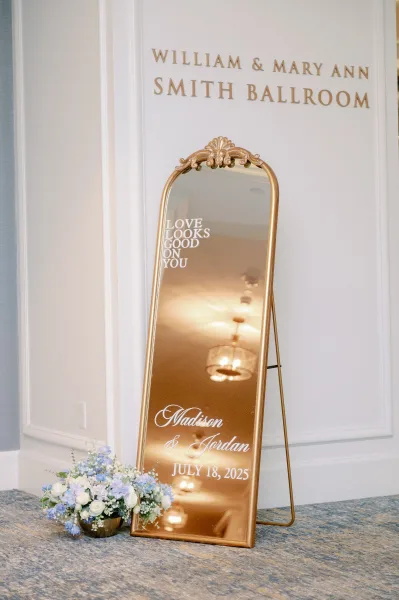 Wedding welcome sign on a gold standing mirror with vinyl lettering and white-and-blue florals in a gold vase at the ballroom entrance