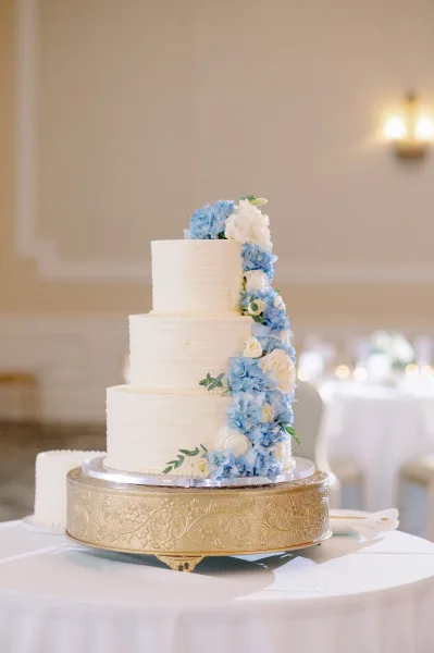 Wedding cake on a gold cake stand, tiered in white buttercream with a blue hydrangea and rose floral cascade in a reception room