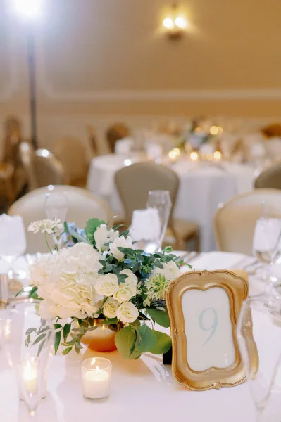 Reception tablescape with wedding table centerpiece of white blooms and greenery, gold table number frame, and votive candles in a banquet hall