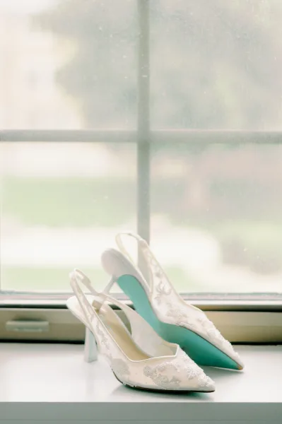 Wedding shoes, bridal heels with white lace slingback pointed toes and teal soles resting on a windowsill in soft window light