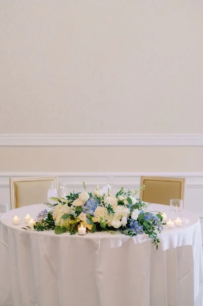Wedding sweetheart table with sweetheart table decor featuring blue hydrangeas, white roses, greenery, and candlelight against a neutral wall