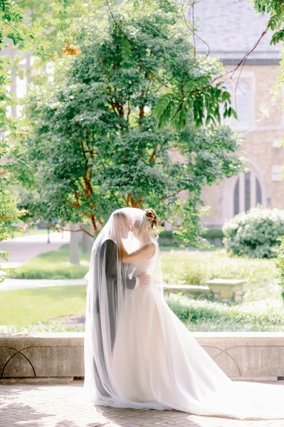 Wedding kiss portrait of bride and groom kissing under a sheer veil, framed by garden trees and a stone wall near a brick building