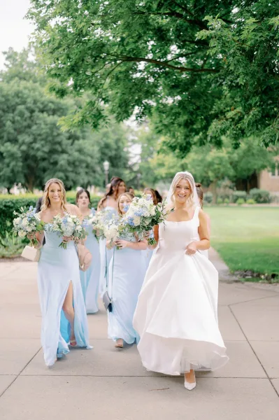 Bridal party portrait of bride with bridesmaids in blue dresses walking under a tree canopy, holding bouquets, bride in veil and satin gown