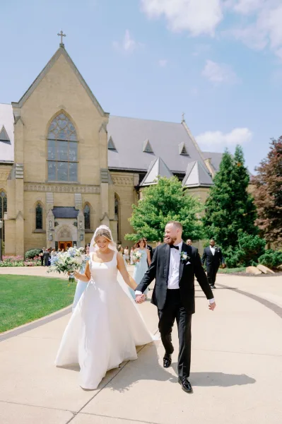 Newlywed couple holding hands, bride in veil with blue and white bouquet, walking from a stone church as bridesmaids follow under blue sky