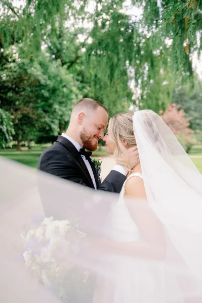 Wedding couple portrait of bride and groom forehead touch, embracing under her veil as she holds a bouquet on a tree-lined park path