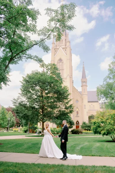 Couple portrait of bride and groom holding hands, walking on a church lawn as she looks back, dress train and bouquet visible under clouds