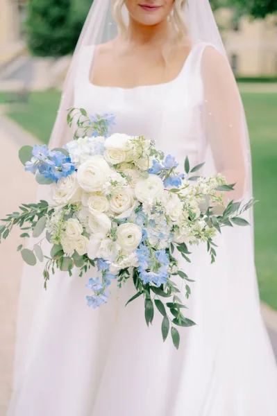 Bridal bouquet of white roses and blue delphinium with eucalyptus greenery, held against a wedding dress and veil on an outdoor walkway