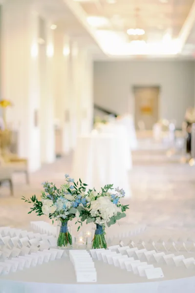 Escort card table with wedding seating cards arranged around floral centerpieces, candles, and greenery in a bright indoor hallway
