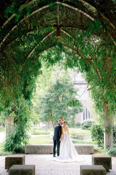 Couple portrait of bride and groom back view holding hands under a pergola with hanging vines, garden lawn and stone building behind