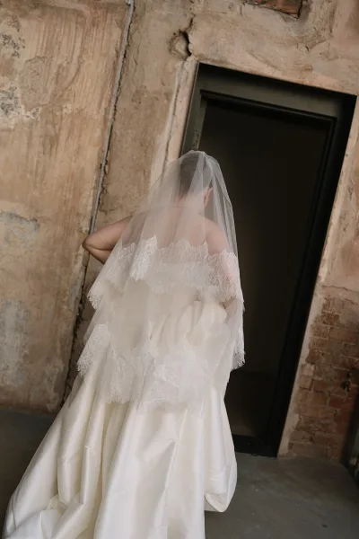 Bridal portrait of a bride walking away, her lace-trimmed veil trailing as she nears a doorway set between a plaster wall and exposed brick.