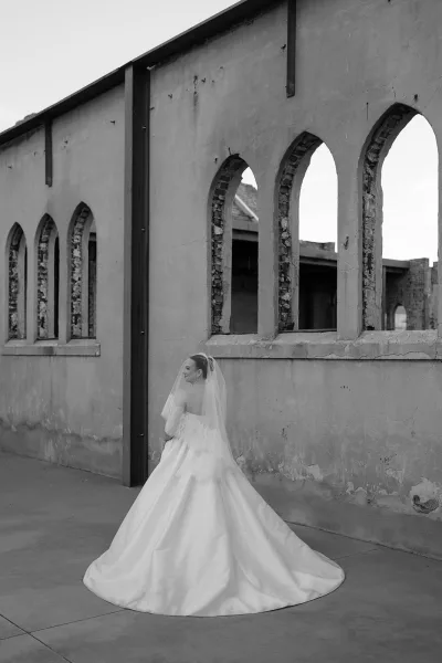 Bridal portrait of a bride in ball gown with a lace bodice, standing by arched windows in industrial ruins against a raw concrete wall.