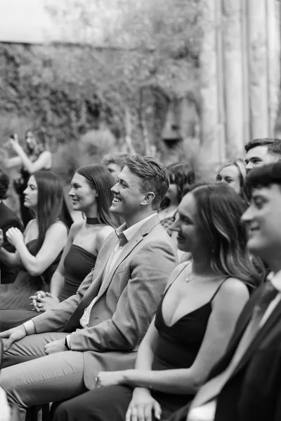 Wedding guests in elegant dresses react with smiles in a candid guest reaction photo, framed by garden foliage and a stone wall.