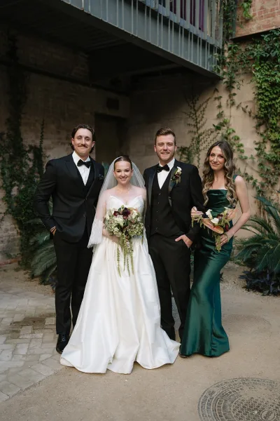 Wedding party portrait of a couple with bridesmaid and groomsman, bride in a classic veil beside an ivy-covered brick wall and metal balcony.