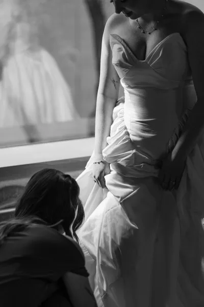 Bride getting ready, adjusting her dress by a window as her reflection shimmers in the glass, a simple necklace glinting at her collarbone.