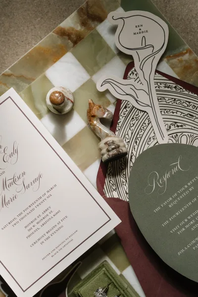 Wedding invitation suite with a sage green wedding invite, styled beside chess pieces on a checkerboard marble slab and stone tabletop.