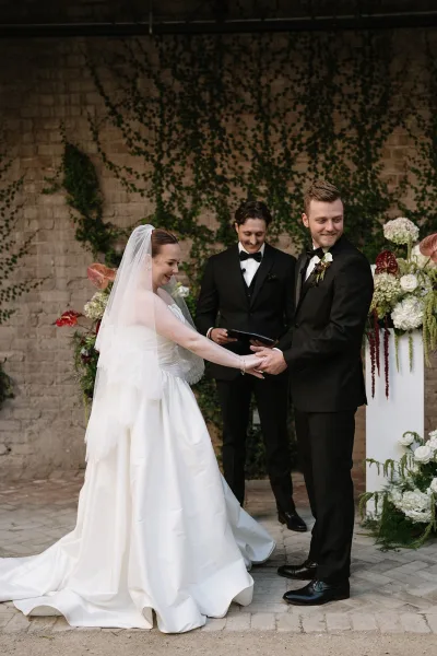 Ceremony moment with a couple holding hands, exchanging vows before a brick wall draped in climbing vines, the bride’s veil catching the light.